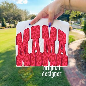 May include: A white t-shirt featuring the word MAMA in large, bold, block letters. The letters have a vibrant pink and red leopard print pattern. Behind the text, there are faint, thin, light gray chevron lines. The text is centered on the shirt.