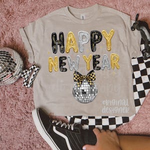May include: Beige t-shirt featuring the text "HAPPY NEW YEAR" in gold, silver, and black bubble letters. A disco ball and a checkered bow are below the text. The words "original designer" are printed below the disco ball.