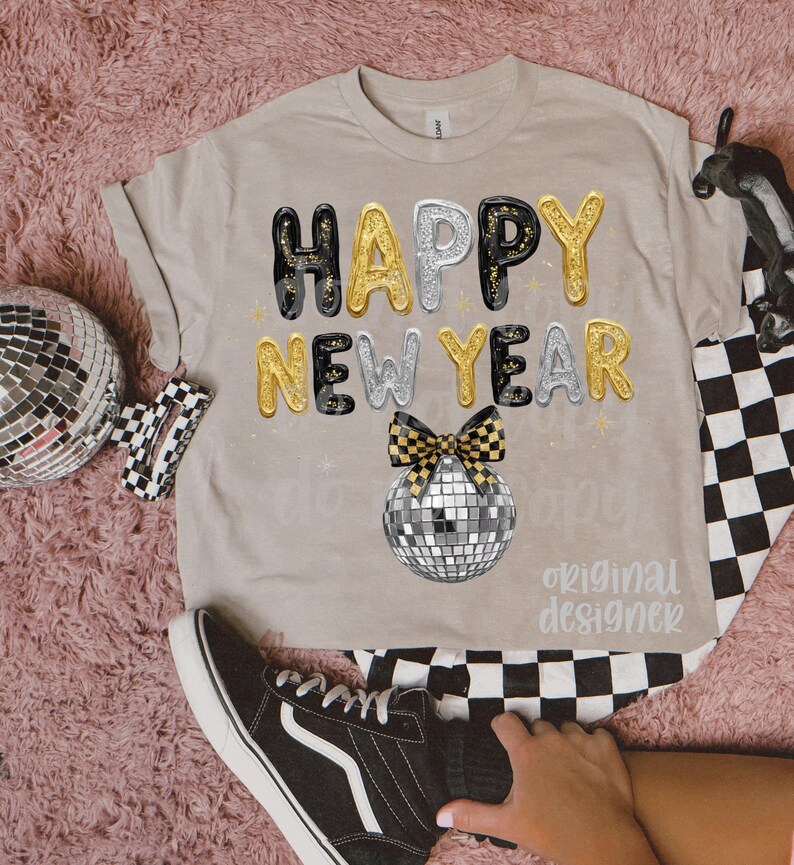 May include: Beige t-shirt featuring the text "HAPPY NEW YEAR" in gold, silver, and black bubble letters. A disco ball and a checkered bow are below the text. The words "original designer" are printed below the disco ball.