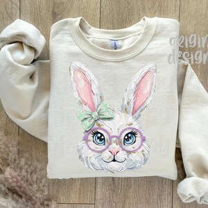 May include: A beige sweatshirt featuring a whimsical illustration of a white bunny wearing glasses and a bow. The bunny has pink-lined ears and blue eyes. The words "original designer" are visible on the top right.