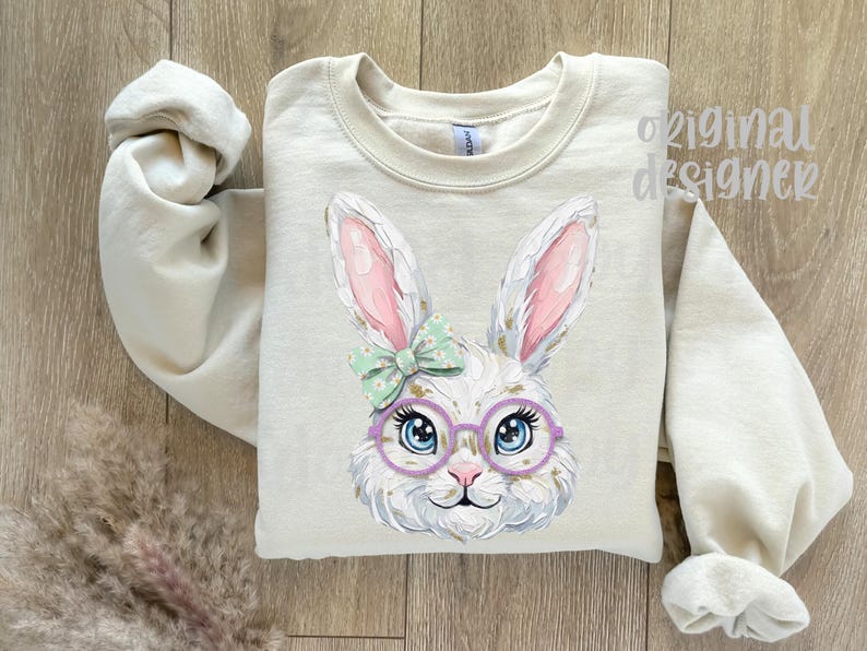 May include: A beige sweatshirt featuring a whimsical illustration of a white bunny wearing glasses and a bow. The bunny has pink-lined ears and blue eyes. The words "original designer" are visible on the top right.