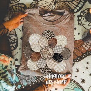 May include: A brown t-shirt with a large, appliqued flower design. The flower petals feature various textures and patterns, including leopard print, gingham, and lace. The text "original designer" is visible.