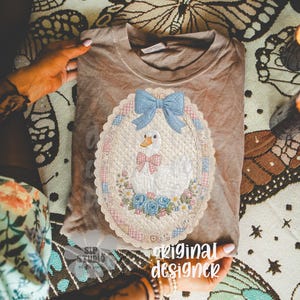 May include: A tan t-shirt featuring an embroidered design of a white goose wearing a pink bow tie, a blue bow, and surrounded by flowers. The design is inside an oval frame with a scalloped edge. The words "original designer" are visible.