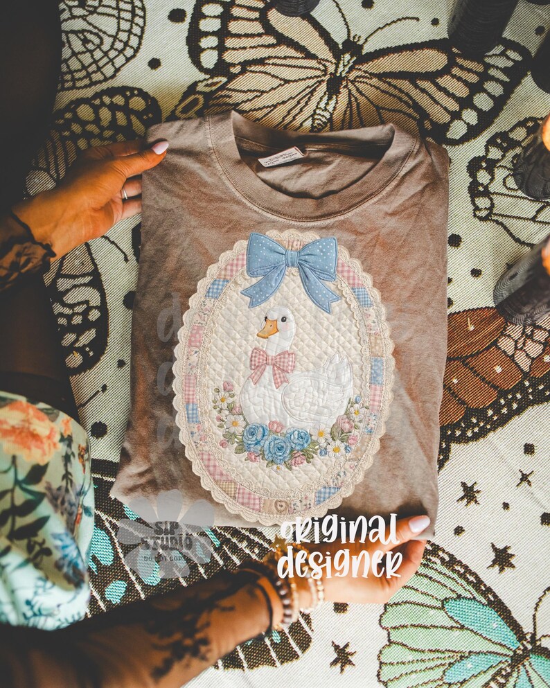 May include: A tan t-shirt featuring an embroidered design of a white goose wearing a pink bow tie, a blue bow, and surrounded by flowers. The design is inside an oval frame with a scalloped edge. The words "original designer" are visible.
