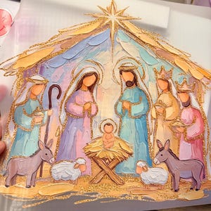 May include: A nativity scene illustration featuring a central star, Mary, Joseph, the infant Jesus in a manger, and three figures, accented with gold glitter outlines. The figures are rendered in pastel blue, pink, and yellow tones with thick paint strokes.