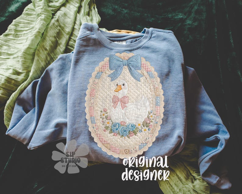 May include: Light blue sweatshirt featuring a quilted design of a white goose with a pink bow, blue bow, and floral accents. The design is set within an oval frame. The text "original designer" is visible.