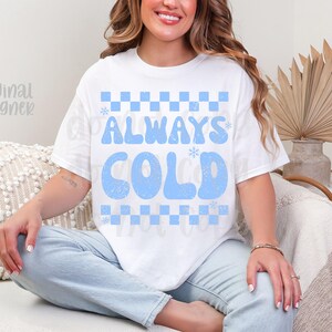 May include: White t-shirt with the text "ALWAYS COLD" in light blue, featuring a checkerboard pattern. The shirt is a casual style, suitable for everyday wear.