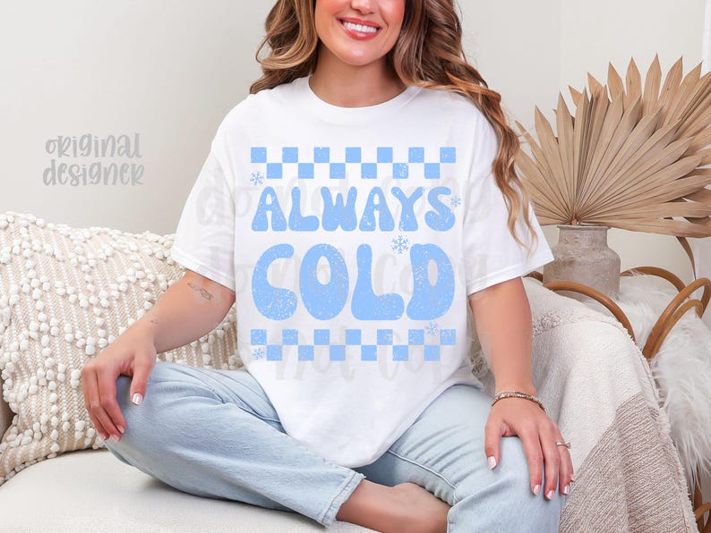 May include: White t-shirt with the text "ALWAYS COLD" in light blue, featuring a checkerboard pattern. The shirt is a casual style, suitable for everyday wear.