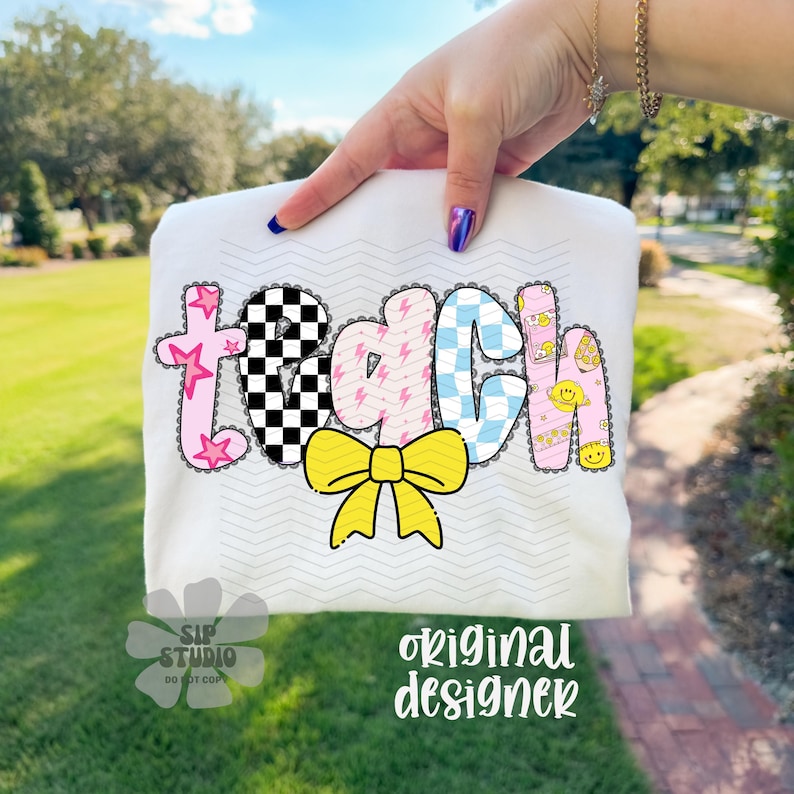 May include: White t-shirt with the word "TEACH" in colorful, patterned letters. The letters feature a checkerboard, lightning bolts, and smiley faces. A yellow bow is below the word. The shirt is held up by a person.