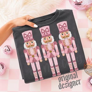 May include: A dark gray sweatshirt featuring a graphic print of three nutcrackers in shades of pink and glittery accents. Each nutcracker wears a tall hat with a bow, a uniform jacket, and striped trousers. The design includes the text Electric Edge Designs.