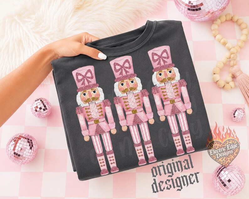 May include: A dark gray sweatshirt featuring a graphic print of three nutcrackers in shades of pink and glittery accents. Each nutcracker wears a tall hat with a bow, a uniform jacket, and striped trousers. The design includes the text Electric Edge Designs.