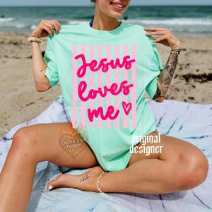 May include: A mint green t-shirt with the text "Jesus loves me" in bright pink, accented with pink and white striped vertical lines. The shirt is a casual, oversized style, perfect for a relaxed day.
