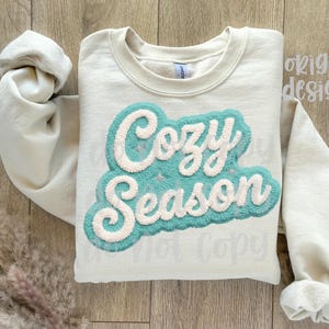 May include: Cream-colored sweatshirt with the words "Cozy Season" in a teal and white textured design. The sweatshirt has long sleeves and a crew neck. The image is taken from a top-down perspective.