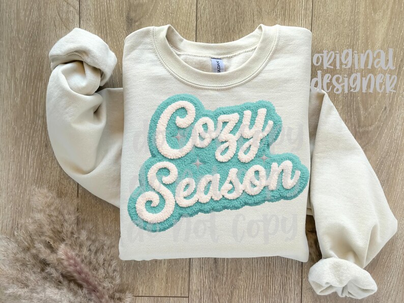 May include: Cream-colored sweatshirt with the words "Cozy Season" in a teal and white textured design. The sweatshirt has long sleeves and a crew neck. The image is taken from a top-down perspective.