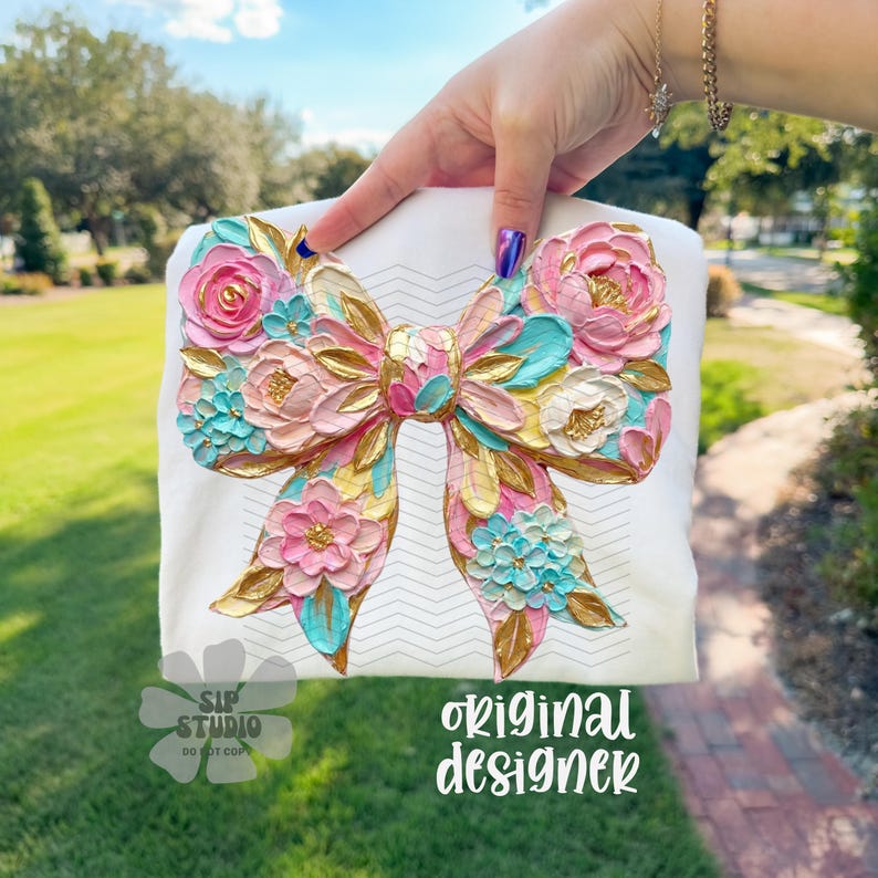 May include: White t-shirt featuring a large, textured bow design. The bow is adorned with pink, gold, and turquoise floral embellishments. The text "original designer" is visible on the shirt. The shirt is held up by a person.