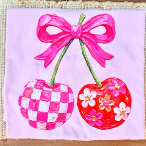 May include: A light pink t-shirt featuring a central graphic of two cherries connected by a bright pink bow. One cherry has a pink and white checkered pattern, while the other is red with white and pink flowers and gold glitter accents.