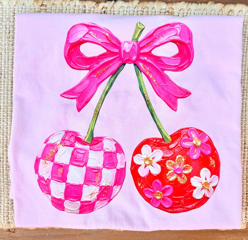 May include: A light pink t-shirt featuring a central graphic of two cherries connected by a bright pink bow. One cherry has a pink and white checkered pattern, while the other is red with white and pink flowers and gold glitter accents.