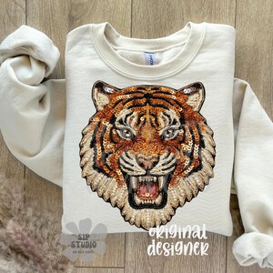 May include: Cream-colored sweatshirt featuring a detailed tiger face design. The tiger's face is intricately embellished with sequins in shades of orange, brown, and gold. The design includes the text "original designer".