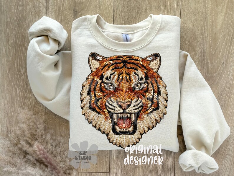 May include: Cream-colored sweatshirt featuring a detailed tiger face design. The tiger's face is intricately embellished with sequins in shades of orange, brown, and gold. The design includes the text "original designer".