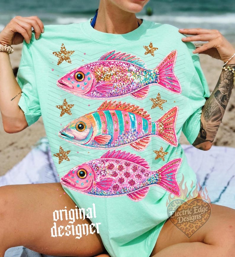 Puede incluir: Camiseta verde menta con tres ilustraciones de peces coloridos, cada uno con purpurina y estrellas doradas. Los peces son rosas, azules y naranjas. La camiseta tiene el texto "original designer" en la parte inferior.