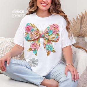 May include: White t-shirt featuring a large, colorful bow design. The bow is adorned with pink, blue, and gold floral accents, creating a textured, 3D effect. The words "original designer" are printed on the shirt.