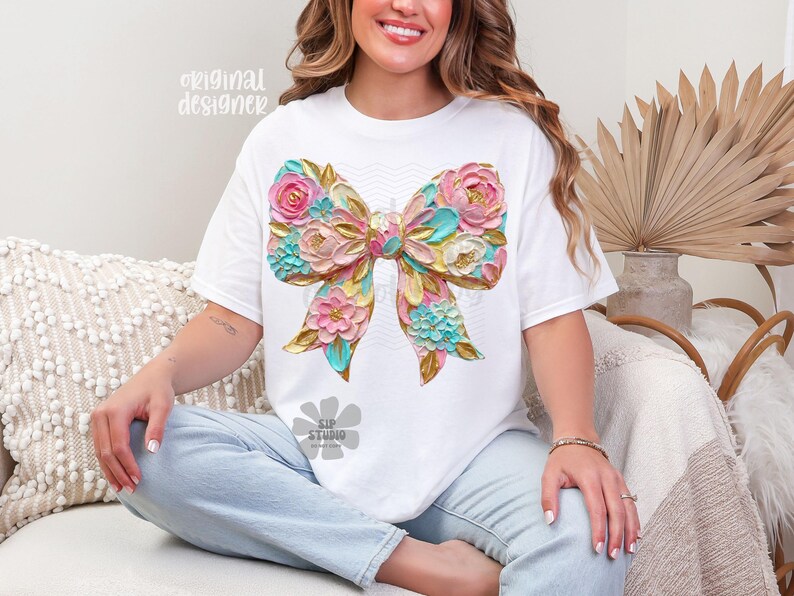 May include: White t-shirt featuring a large, colorful bow design. The bow is adorned with pink, blue, and gold floral accents, creating a textured, 3D effect. The words "original designer" are printed on the shirt.