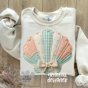 May include: Cream-coloured sweatshirt with a seashell design. The seashell is made of pastel-coloured fabrics, including pink, blue, and white, with pearl accents and a lace bow. The words "original designer" are printed on the bottom.