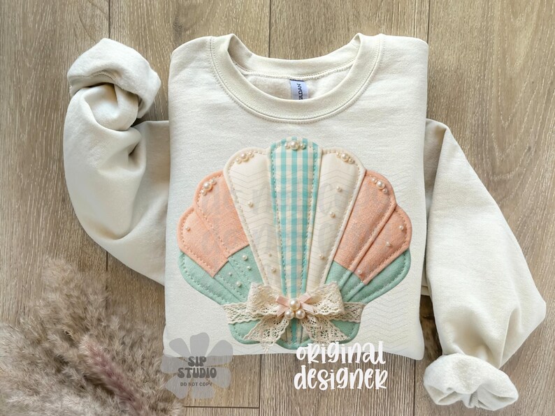 May include: Cream-coloured sweatshirt with a seashell design. The seashell is made of pastel-coloured fabrics, including pink, blue, and white, with pearl accents and a lace bow. The words "original designer" are printed on the bottom.