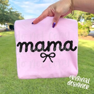 May include: A light pink sweatshirt with the word "mama" in black, rope-like lettering and a black bow. The words "original designer" are in white script in the bottom right corner. The sweatshirt is held up by a hand with purple nail polish.