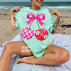 May include: A mint green t-shirt featuring a large graphic of two cherries connected by a bright pink bow. One cherry has a pink and white checkered pattern, while the other is solid red with white and yellow flowers. The text original designer is visible.