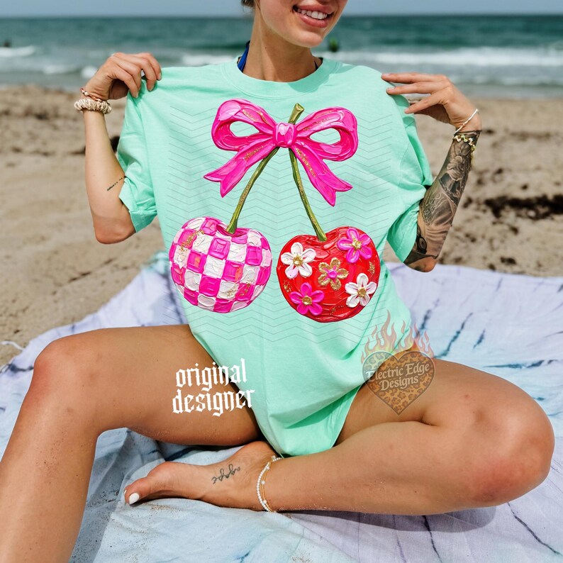 May include: A mint green t-shirt featuring a large graphic of two cherries connected by a bright pink bow. One cherry has a pink and white checkered pattern, while the other is solid red with white and yellow flowers. The text original designer is visible.