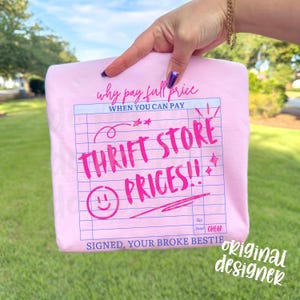 May include: A light pink t-shirt with the text "Why pay full price when you can pay Thrift Store Prices!!" in a handwritten style. The design resembles a receipt with a smiley face and stars. The words "Signed, Your Broke Bestie" are also included.