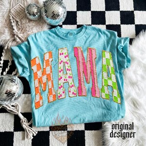 May include: A light blue t-shirt with the word "MAMA" in large, colorful letters. The "M" is orange and white checkered, the first "A" is pink with cherry designs, the second "M" is pink, and the second "A" is green and white checkered.