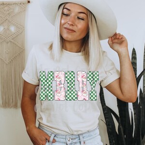 May include: Cream-colored t-shirt with the word "MERRY" in a block letter design. The letters are a mix of green and white checkered patterns and a pink floral pattern with Santa Claus illustrations. The shirt is a casual style.