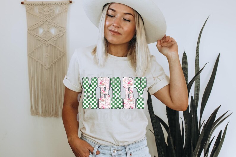 May include: Cream-colored t-shirt with the word "MERRY" in a block letter design. The letters are a mix of green and white checkered patterns and a pink floral pattern with Santa Claus illustrations. The shirt is a casual style.