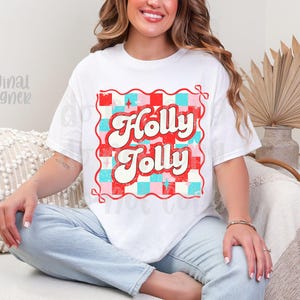 May include: White t-shirt with a retro design that says "Holly Jolly" in red and white, set against a red, pink, and blue checkered background. The shirt has a vintage, distressed look.