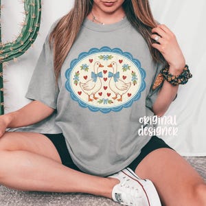 May include: Gray t-shirt featuring a whimsical design of two geese with blue bows, surrounded by hearts and floral accents within a blue scalloped oval. The text "original designer" is printed on the shirt.