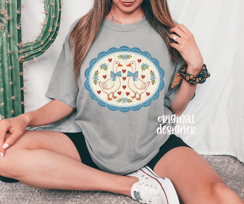 May include: Gray t-shirt featuring a whimsical design of two geese with blue bows, surrounded by hearts and floral accents within a blue scalloped oval. The text "original designer" is printed on the shirt.