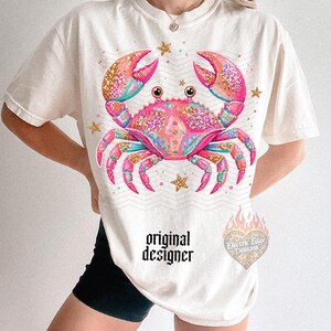 May include: Cream-colored t-shirt featuring a vibrant crab design with pink, teal, and gold glitter accents. The crab is surrounded by gold stars. The text "original designer" is printed below the crab.