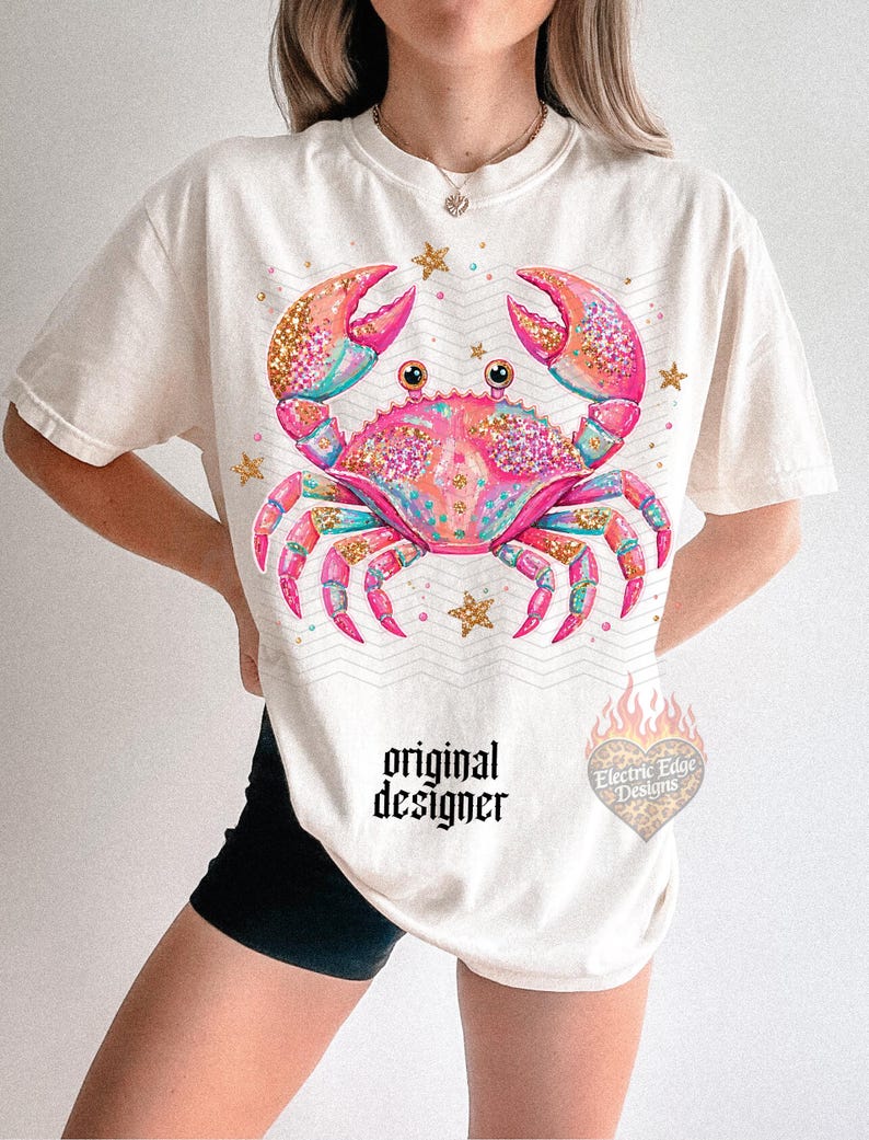May include: Cream-colored t-shirt featuring a vibrant crab design with pink, teal, and gold glitter accents. The crab is surrounded by gold stars. The text "original designer" is printed below the crab.