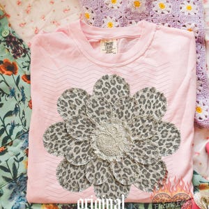 May include: A light pink Comfort Colors t-shirt featuring a large flower design made of leopard print fabric and a lace center. The words "original designer" are printed in a gothic font. The shirt is folded on a floral patterned surface.