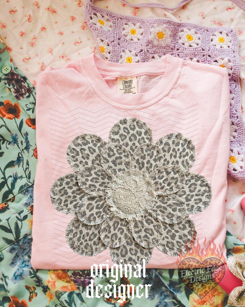 May include: A light pink Comfort Colors t-shirt featuring a large flower design made of leopard print fabric and a lace center. The words "original designer" are printed in a gothic font. The shirt is folded on a floral patterned surface.
