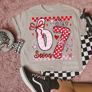 May include: A beige t-shirt featuring the numbers "6" and "7" in a decorative design with hearts and bows. The word "Six" is above the "6", and "Seven" is below. The shirt has a checkerboard pattern and the words "original designer".
