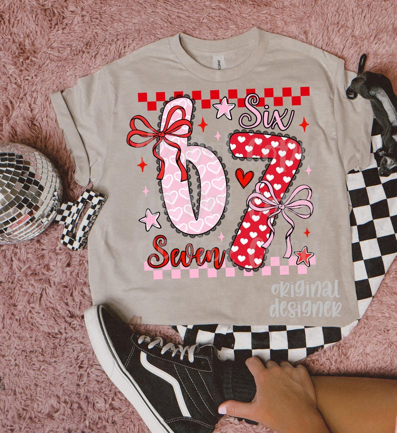 May include: A beige t-shirt featuring the numbers "6" and "7" in a decorative design with hearts and bows. The word "Six" is above the "6", and "Seven" is below. The shirt has a checkerboard pattern and the words "original designer".