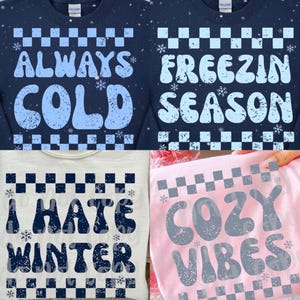May include: Four sweatshirts with winter-themed text. Two navy blue sweatshirts say "ALWAYS COLD" and "FREEZIN SEASON" in light blue. A light gray sweatshirt says "I HATE WINTER" in navy blue. A pink sweatshirt says "COZY VIBES" in gray.