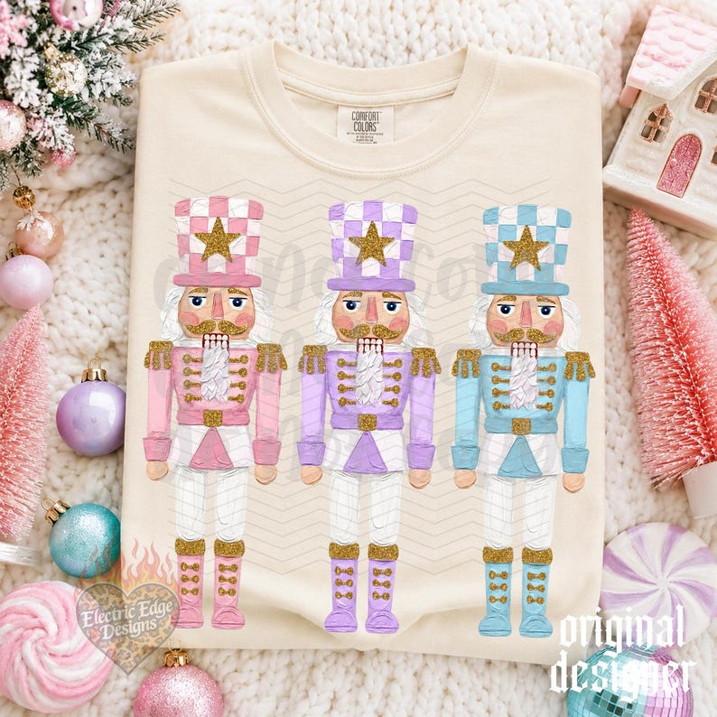 May include: A cream-colored t-shirt featuring a graphic print of three nutcracker figures in pastel pink, lavender, and light blue. Each nutcracker wears a tall hat with a gold star, a matching uniform with gold accents, and gold boots.