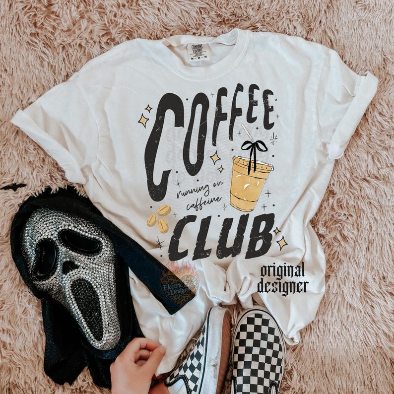 May include: Cream-colored t-shirt with the words "COFFEE CLUB" in black, featuring a coffee cup graphic and the phrase "running on caffeine." The shirt also has a black and silver mask and checkered shoes.