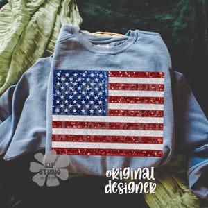 May include: A light blue sweatshirt featuring a sequined American flag design. The flag has a blue field with white stars and red and white stripes. The words "original designer" are written in white script.