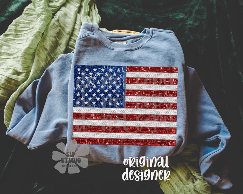 May include: A light blue sweatshirt featuring a sequined American flag design. The flag has a blue field with white stars and red and white stripes. The words "original designer" are written in white script.