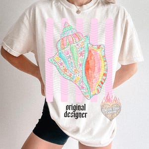 May include: Cream-colored t-shirt featuring a colorful seashell design with pink vertical stripes. The seashell is detailed with various sea creatures and vibrant colors. The text "original designer" is printed below the seashell.